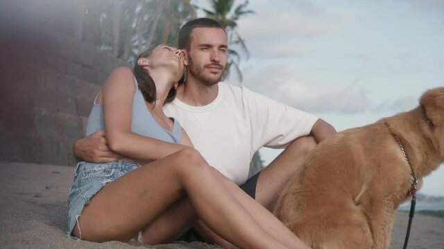 Side view medium long of dark-haired beautiful Caucasian girl hugging and kissing her young boyfriend in white t-shirt on sunny beach. Romantic couple with dog