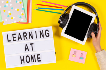 Learning at home. Hand holding digital tablet computer with headphones, lightbox, school supplies on yellow background. Top view