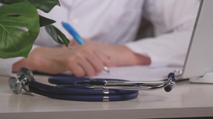 Close up view of stethoscope medical equipment with female doctor working in background. Professional physician gp wearing white coat writing notes at desk. General practice concept