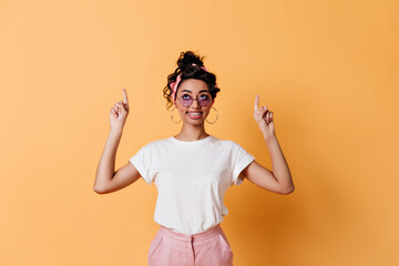 Glad mixed race girl pointing with fingers. Studio shot of good-humoured lady in sunglasses gesturing on yellow background.