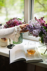 a woman's hand reaches for a bouquet of lilacs by the window