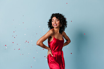 Appealing mixed race girl standing under confetti. Studio shot of laughing curly woman in red dress.