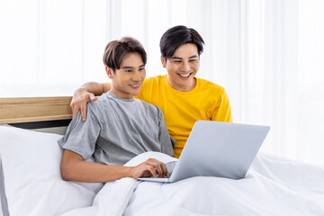 Happy Asian homosexual gay couple sitting on white bed using laptop computer together, LGBT concept.