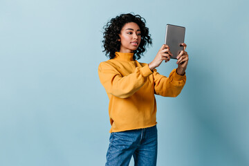 Mixed race girl holding digital tablet. Studio shot of curious lady with gadget isolated on blue...