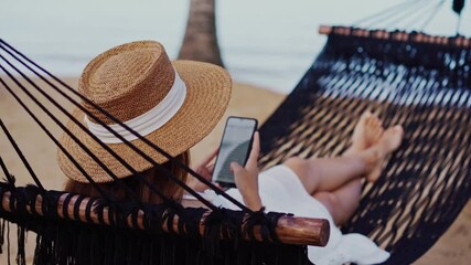 Young woman traveler lying on a hammock and using smartphone at the beach while traveling for summer vacation	
