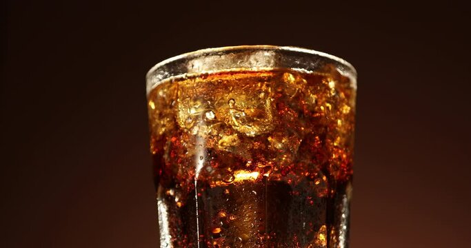 Cola glass with ice cubes slowly rotating close-up. Cold soda drink spinning over brown background. 4K video footage