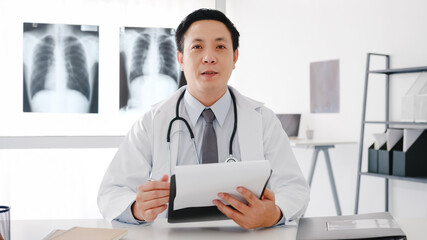 Young Asia male doctor in white medical uniform with stethoscope using computer laptop talk video...