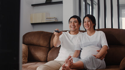 Young Asian Pregnant couple watching TV. Mom and Dad feeling happy smiling peaceful while take care baby, pregnancy lying on sofa in living room at home concept.