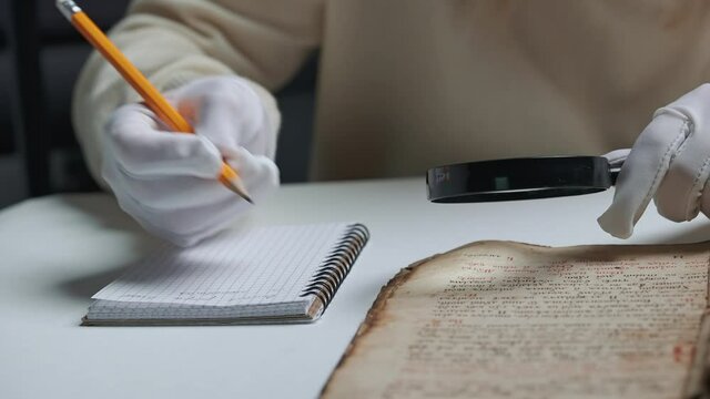 Researcher in cotton gloves examines antique book with magnifying glass. Scientific translation of ancient literature. Studying manuscript with ancient writings
