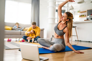Young woman, mother exercising at home in living room, father playing with kids in background.