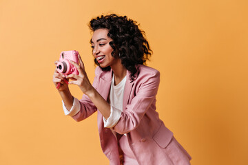 Smiling woman in pink jacket taking pictures. Studio shot of trendy mixed race girl with camera.