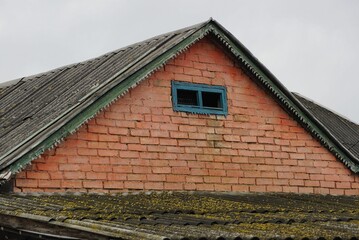 old red brick loft of a rural house with a small blue window under a gray slate roof against the sky