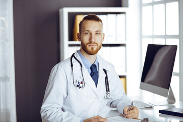 Red-bearded doctor standing straight in clinic near his working place. Portrait of physician. Medicine and healthcare concept