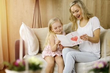 Happy mother day in sunny flat. Child daughter congratulates mom and gives her postcard with heart...