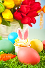 Fototapeta na wymiar Easter funny bunny cupcake with colorful eggs. Easter celebration festive table. Basket of flowers tulips on the background.