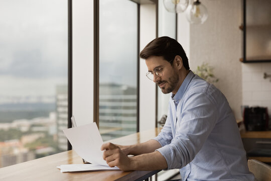 Successful businessman read paper document at work desk in studio apartment near panoramic window. Thoughtful confident young man freelancer engaged in paperwork at home office study official letter