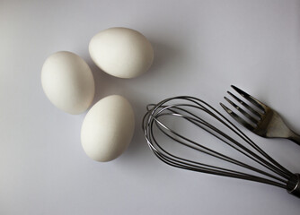 three chicken white eggs whisk and fork on gray background