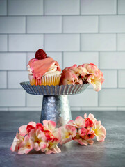 Raspberry cupcakes with white frosting swirled high with a pink drizzle and a fresh raspberry on top on a raised rustic metal cake display dish with a gray counter and white subway tile background.