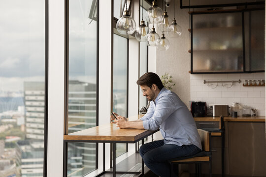 Smiling young bearded man sit at kitchen desk near big panoramic window relax surf internet using smartphone. Happy millennial guy spend leisure time in social network shopping chatting on modern cell