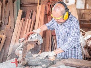 Elderly carpenter craftsman wearing safety gear working with hardwood using a miter saw to cut a...
