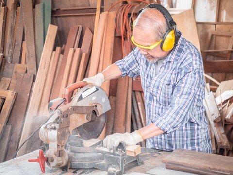 Elderly carpenter craftsman wearing safety gear working with hardwood using a miter saw to cut a piece of wood in the workshop. Manufacture of wood products.. - Powered by Adobe