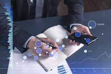 A trader in formal wear writing down some quotes to research stock market trends using smart phone for right investment solutions. Wealth management concept. Hologram Forex chart over close up shot.