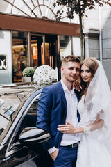 A romantic walk of the bride and groom through the city streets. Newlyweds on the background of a black car.