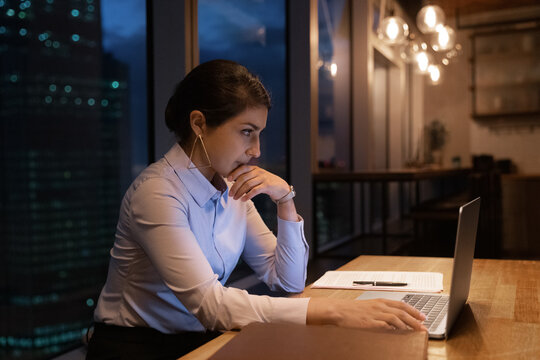 Thoughtful Indian Businesswoman Work By Laptop At Studio Apartment Or Modern Office While Night Cover City Outside Panoramic Window. Pensive Young Hindu Female Read Document Online On Computer Screen