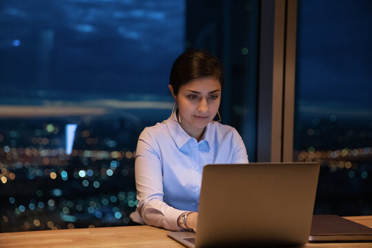 Taken with work. Concentrated indian woman focused on pc screen sitting at table by picture window with evening urban lights outside. Young mixed race lady employee type email on laptop by office desk