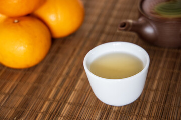 A cup of hot Japanese green tea in a white ceramic teacup put on bamboo sheet with clay porcelain teapot and mandarin orange.