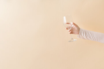 Female hand holding empty clean transparent wine glass against beige background.Copy space, minimal composition.