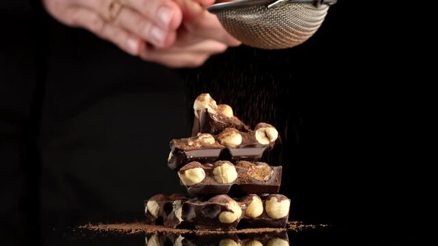 sprinkle chocolate bar with cocoa powder. stack of dark chocolate with nuts on black background. confectionery concept