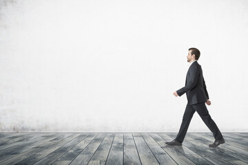 Handsome businessman walking in minimalistic interior with blank concrete wall and wooden floor. Mock up