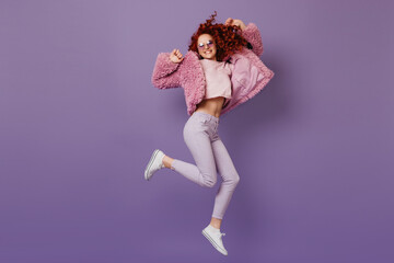 Positive redhead girl in eco-coat, pink top and white pants jumping on lilac background