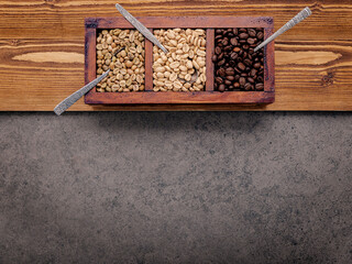 Green and brown unroasted and dark roasted coffee beans in wooden box with spoons setup on dark concrete background.