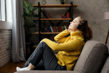 Obraz premium Beautiful pleased woman listening to music in headphones while sitting on sofa at home