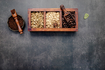 Green and brown unroasted and dark roasted coffee beans in wooden box with scoops setup on dark concrete background.