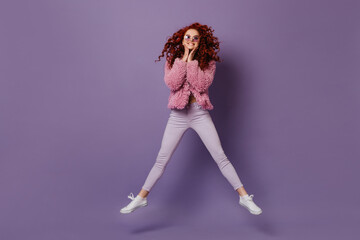 Fototapeta na wymiar Smiling girl in round sunglasses jumps. Woman with red curls in bright outfit is having fun on purple background