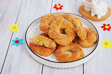 Traditional buttery twisted Italian Easter cookies with multicolored sugar decor on a white plate on a white wooden background. Easter sweets.