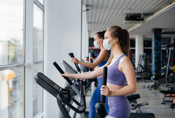 Fototapeta premium Two young beautiful girls exercise in the gym wearing masks during the pandemic. Social distancing in public places.