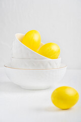 Boiled eggs dyed or painted in vibrant yellow color laying in porcelain bowls standing one upon the other prepared for easter celebration party on white wooden table at kitchen. Vertical image