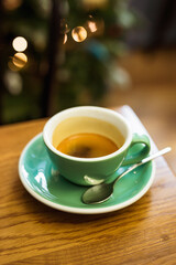 A vertical lifestyle photo of a green cup of black coffee on a saucer, a spoon, bokeh lights in the background, selective focus