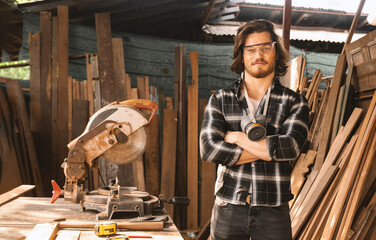 Portrait of a carpenter wearing googles and dust mask standing and crossed arms with table woodwork tool at sawmill.