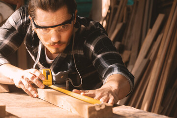 Carpenter wearing googles using a tape measure to measure and check wood plank at sawmill.