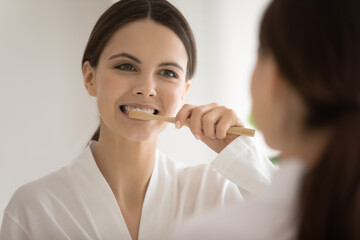 Happy millennial Caucasian woman hold eco wooden toothbrush clean teeth in home bathroom. Smiling young female use bamboo ecological brush for dental oral routine. Dentistry, fresh breath concept.