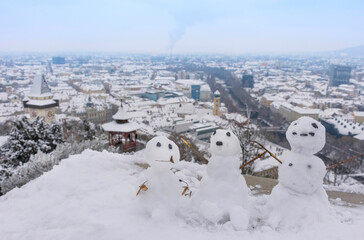 Three funny snowman and the famous clock tower on Schlossberg hill in the background, with snow, in Graz, Styria region, Austria, in winter day. Selective focus