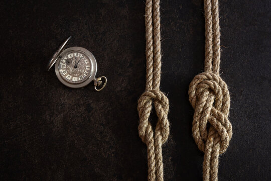 Flat lay composition on a dark background of a steel vintage pocket watch with an open lid and two parallel ropes with tied nautical knots.