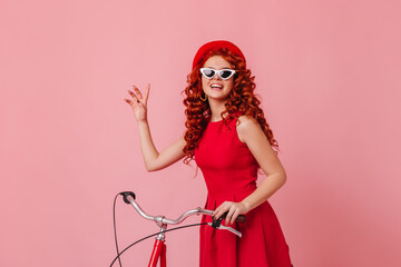 Positive redhead lady in red fashionable outfit and white-rimmed glasses posing with smile on pink background with bicycle
