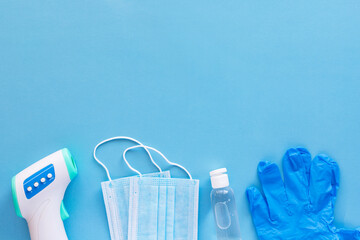 Corona virus protection concept flat lay. Non-contact infrared body thermometer, latex gloves, disposable masks and sanitizer on blue background with copy space for text.