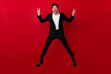 Funny man in suit posing emotionally on red background. Studio shot of caucasian boy jumping with smile.